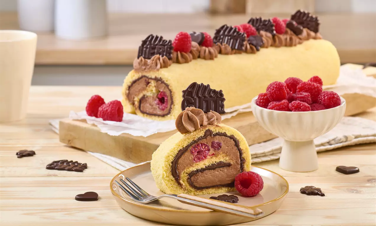 Biskuitrolle mit Schokocreme und Himbeeren, dekoriert mit Schoko-Botschaft u Sahnetupfen. Ein Stück liegt auf einem Teller mit Gabel, daneben eine Schale frischer Himbeeren und verstreute Schokodekor