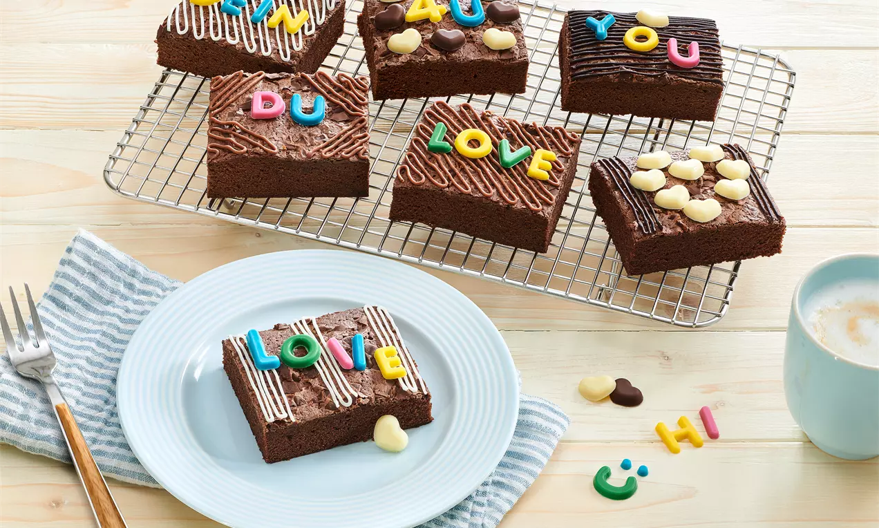Schokoladige Brownie-Stücke mit bunter Zuckerdekor-Beschriftung und cremeweißen Schokoladenherzen auf weißem Teller und Gitter