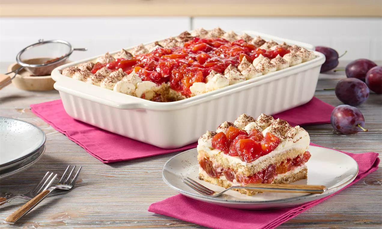 Zwetschgen-Tiramisu in weißer Auflaufform mit Schichten aus Sahnecreme, Löffelbiskuits, Zwetschgen und Kakao, angerichtet auf pinkem Stoff