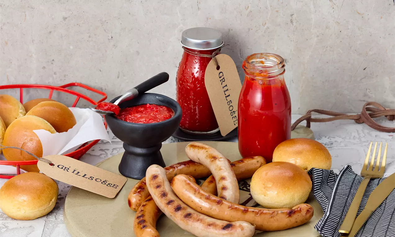 Geräucherte Bratwürste auf einem hellen Teller, mit weichen goldbraunen Brötchen, roter Grillsoße in Glasgefäßen und einer schwarzen Schale