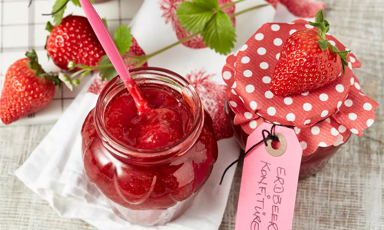 Leuchtend rote Erdbeer-Konfitüre in einem Glas mit rosa Löffel, daneben ein Marmeladenglas mit gepunktetem Stoffdeckel und Erdbeeren