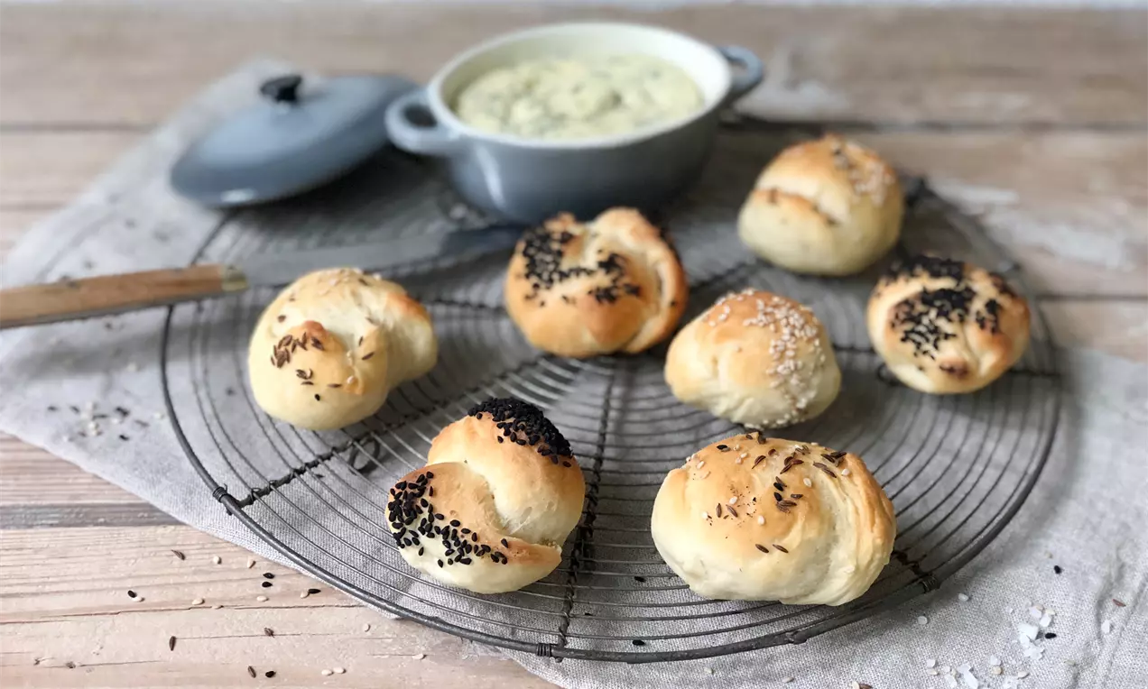 Goldbraune Quark-Öl-Teig-Brötchen mit schwarzem und hellem Sesam bestreut auf einem runden Gitter neben einer grauen Schüssel mit Dip