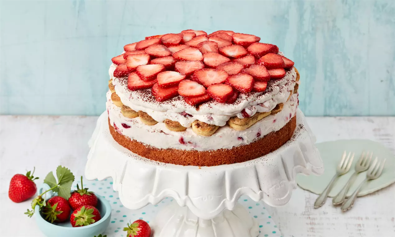 Erdbeer-Tiramisu-Torte mit goldbraunem Boden, heller Creme und frischen Erdbeerscheiben dicht aneinandergereiht auf weißer Tortenplatte
