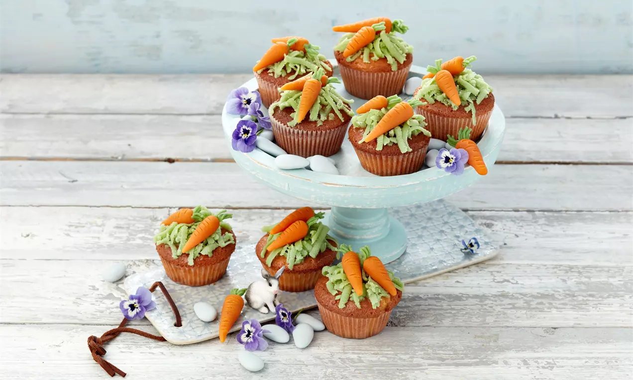 Rübli-Muffins mit orangenen Marzipanmöhren und grünem Frosting auf einer weißen Tortenplatte dekoriert mit Blüten und Zuckersteinen