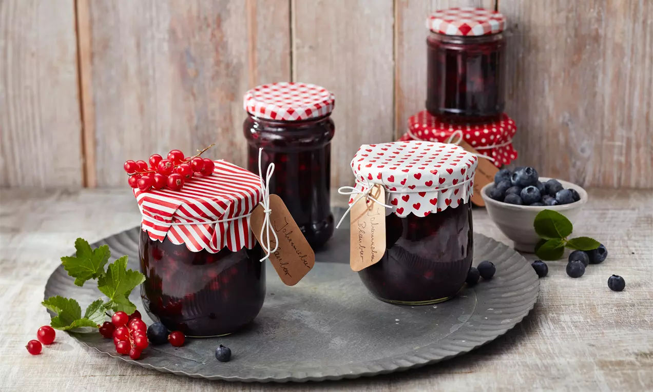 Gläser mit dunkelroter Konfitüre, verziert mit rot gemusterten Deckeln und Anhängern, arrangiert auf einem Zinnteller mit Beeren und Blättern
