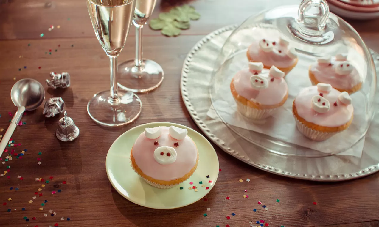 Zartrosa verzierte Glücksschwein-Cupcakes mit kleinen Ohren und Nasen, arrangiert unter einer Glasglocke und auf einem weißen Teller