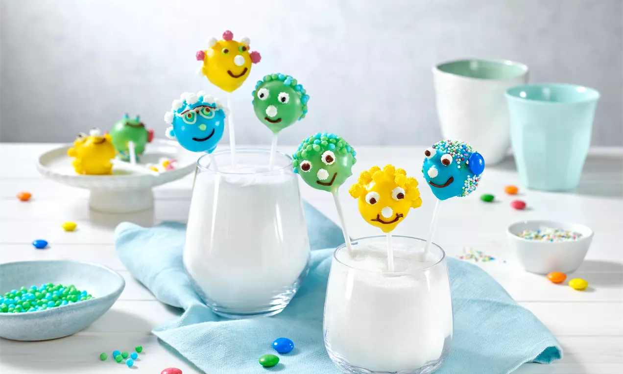 Bunte Cake-Pops mit Gesichtern und Streuseln, auf weißen Gläsern mit Zucker arrangiert, umgeben von bunten Perlen und hellem Geschirr