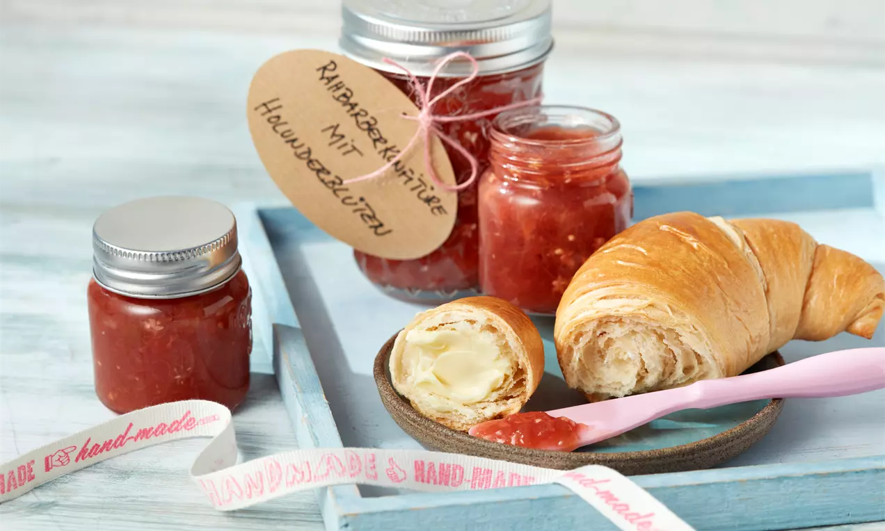 Rhabarberkonfitüre mit rötlich-glänzender Textur in Gläsern, dekoriert mit handbeschriftetem Anhänger neben Croissant und rosa Messer