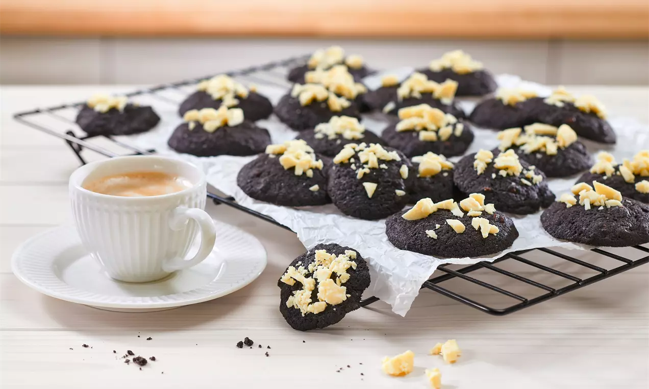 Weiche, tiefschwarze Cookies mit heller, krümeliger Dekoration auf weißem Backpapier und schwarzem Gitter neben einer Tasse Kaffee