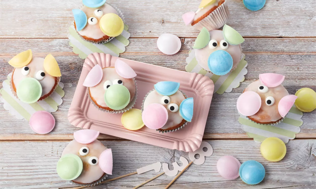 Glücksschweinchen aus rosafarbenem Zuckerguss mit bunten Ohren und runden Nasen auf pastellfarbenem Tablett und Holzuntergrund liegend