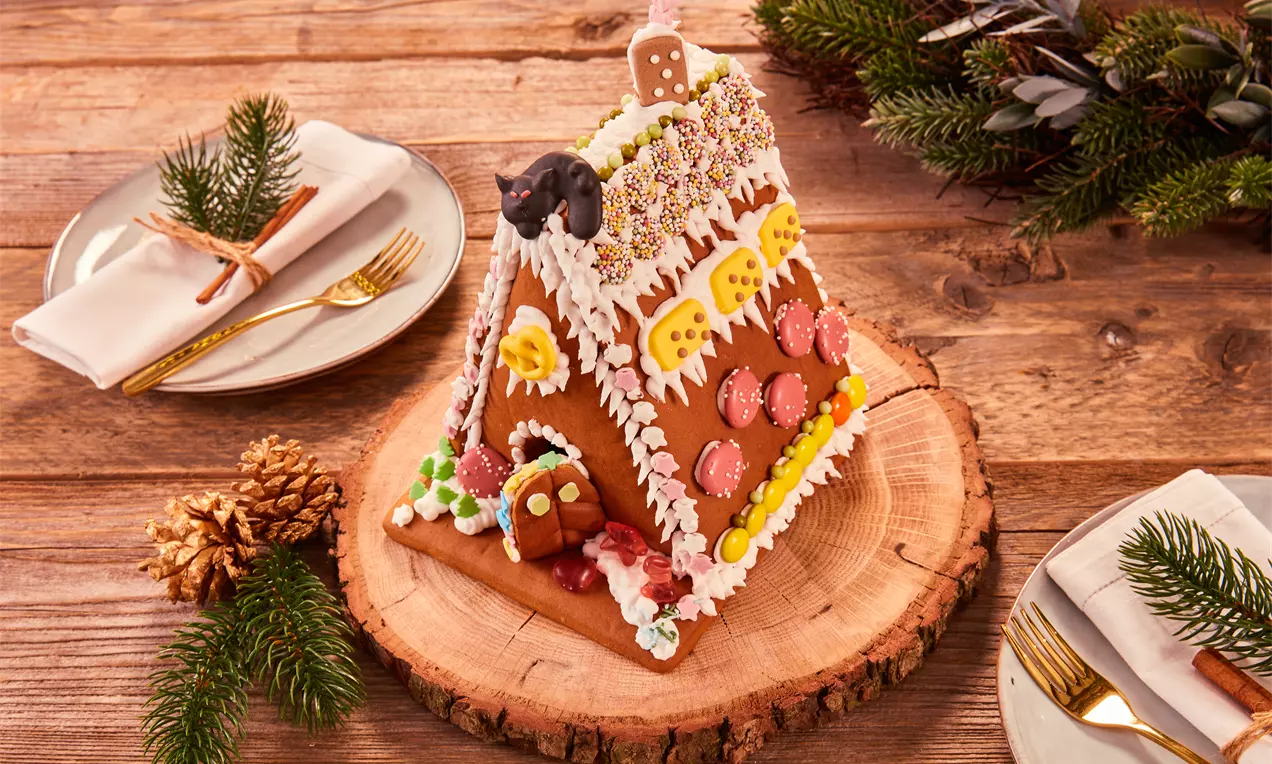 Knusperhäuschen aus braunen Lebkuchenplatten mit bunter Zuckerdeko auf einer Holzscheibe, umgeben von Tannenzweigen und goldenen Bestecken