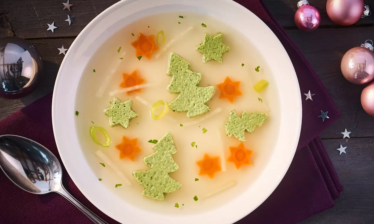 Klare Brühe mit grünen Broteinlagen in Form von Tannenbäumen, orangefarbenen Möhrensternen und feinen Lauchringen auf weißem Teller