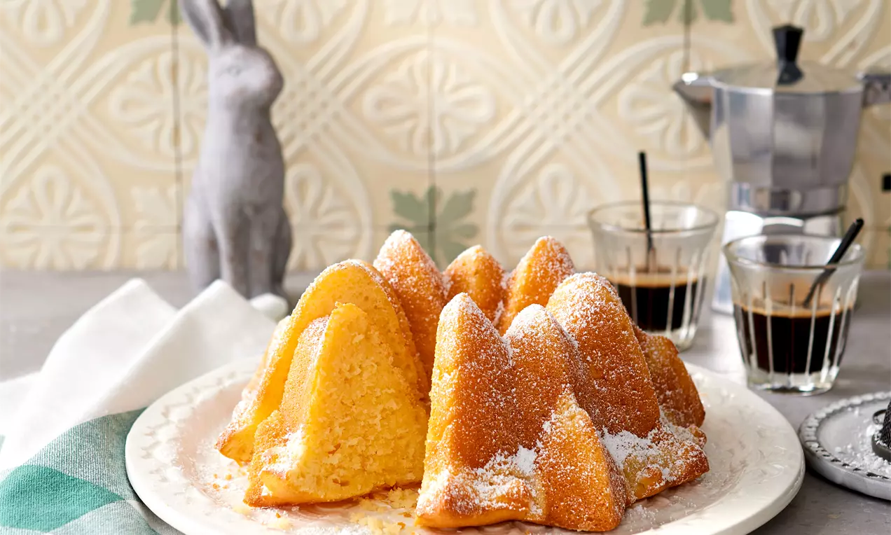 Goldgelber Ciambella in zarter Kranzform, mit weichem Anschnitt und Puderzucker bestäubt auf weißem Teller neben Kaffee und Deko-Elementen