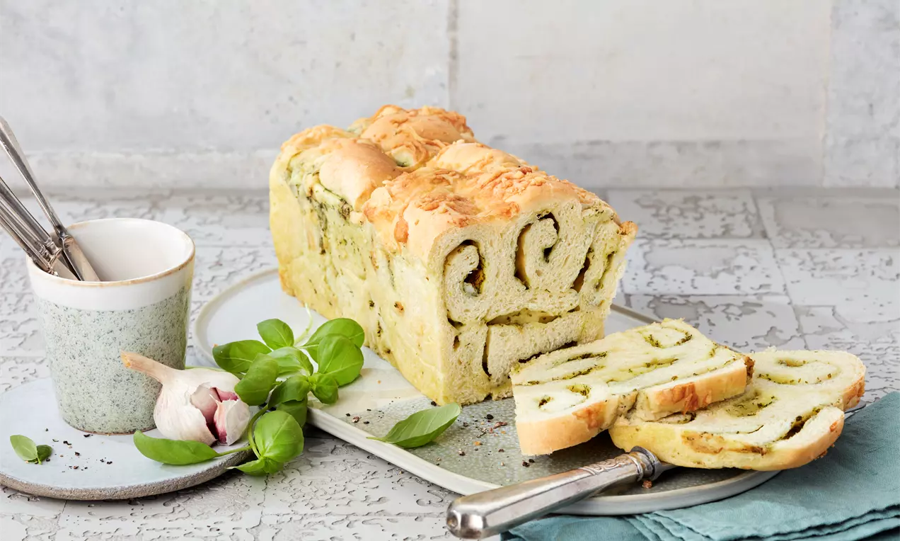 Goldbraunes Zupfbrot mit grüner Pesto-Marmorierung, teils aufgeschnitten und angerichtet auf weißem Teller mit Basilikum und Knoblauchknolle