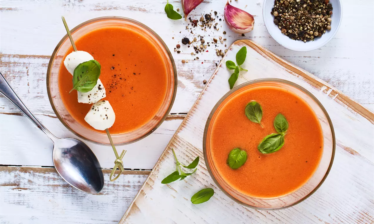 Tomatensuppe in zwei Schalen mit glatter, leuchtend roter Oberfläche, garniert mit frischem Basilikum, daneben Mozzarella-Spieß und Pfefferkörner