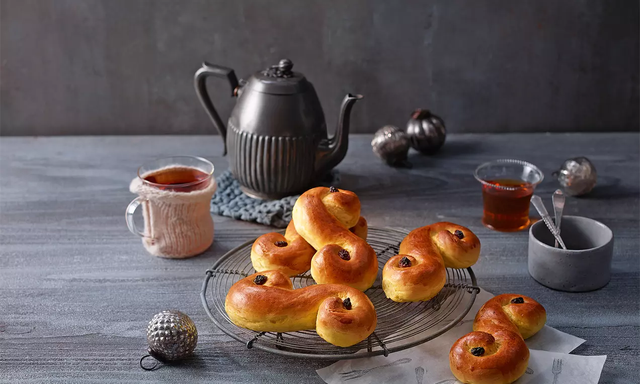 Goldgelb glänzende Lussekatter mit geschwungener S-Form und dunklen Rosinen, leicht versetzt auf einem runden Metallgitter mit Teekanne im Hintergrund