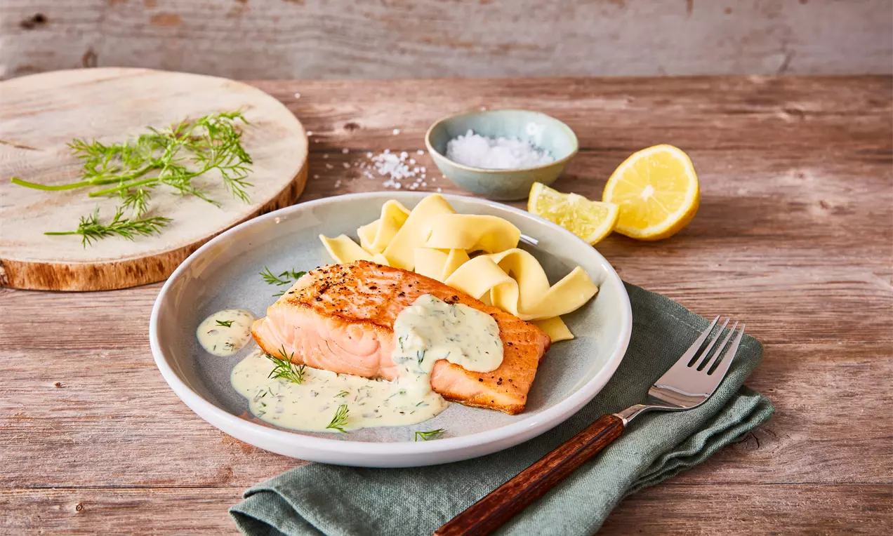 Lachsfilet mit knuspriger Haut auf weißem Teller, umgeben von cremiger Zitronen-Dill-Soße, Zitronenscheibe und Dillzweig