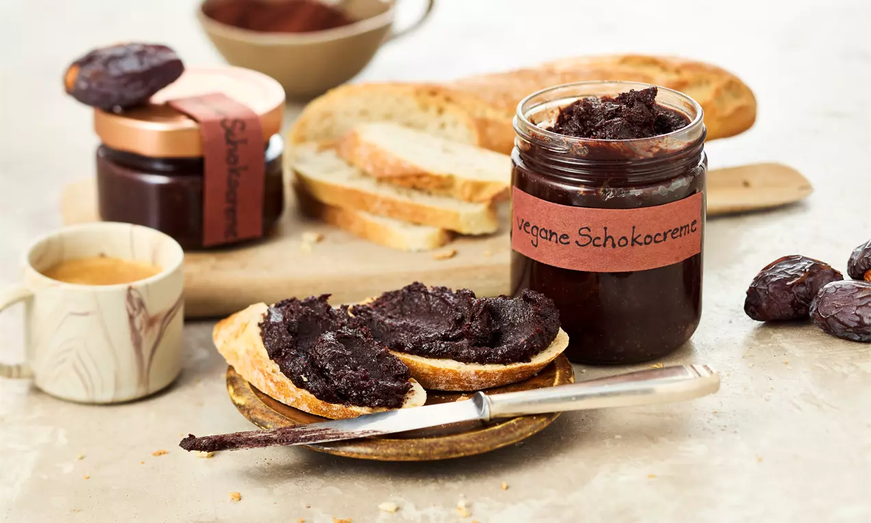 Frühstücksszene mit aufgeschnittenem Brot, bestrichen mit dunkler veganer Schokocreme aus einem Glas. Daneben ein Messer, Datteln, eine Tasse Kaffee und ein weiteres Glas mit Aufstrich.