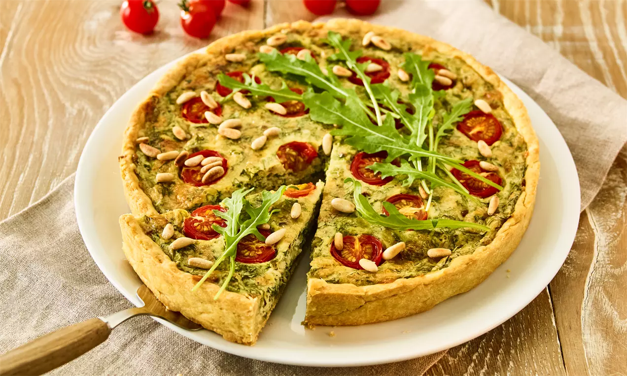 Goldbraune Rucola-Käse-Quiche mit grüner Füllung, roten Tomaten, gerösteten Pinienkernen und frischem Rucola auf weißem Teller