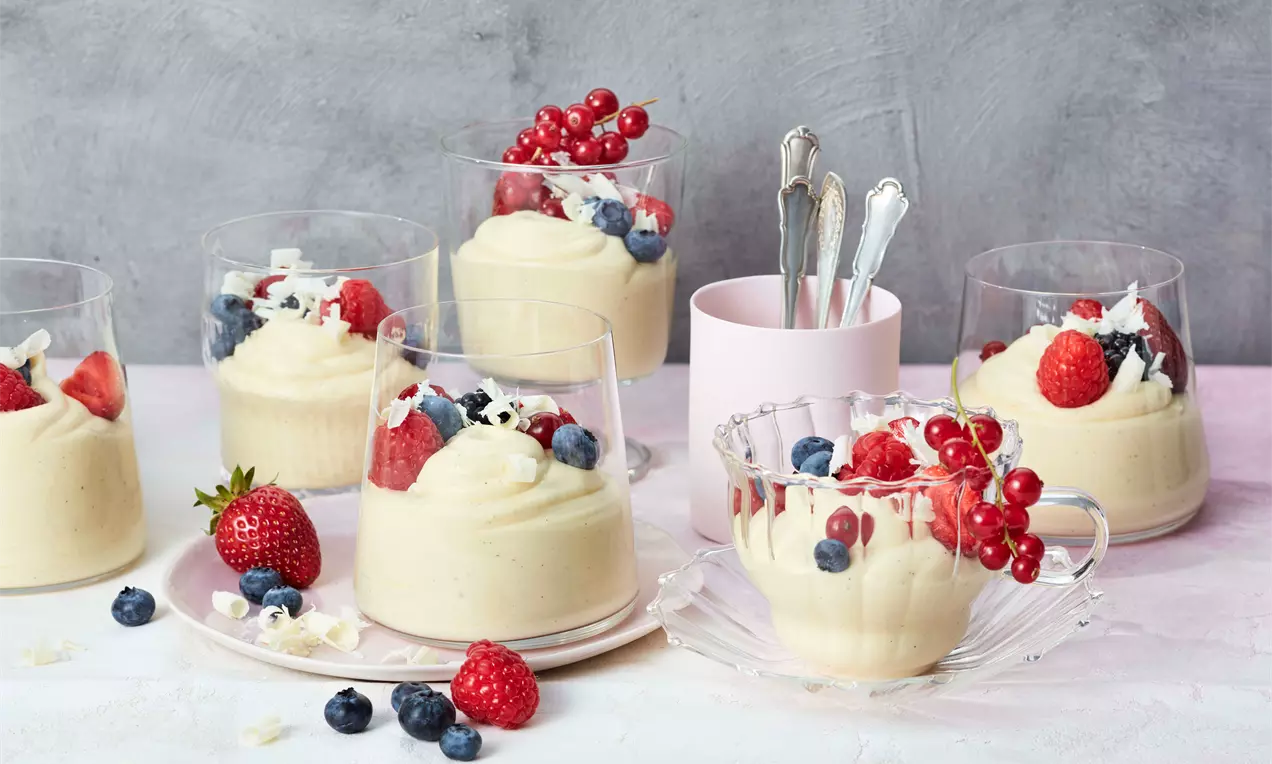 Weiße Mousse au Chocolat cremig serviert in klaren Gläsern, dekoriert mit roten Beeren, Blaubeeren und Schokoraspeln auf hellem Tisch