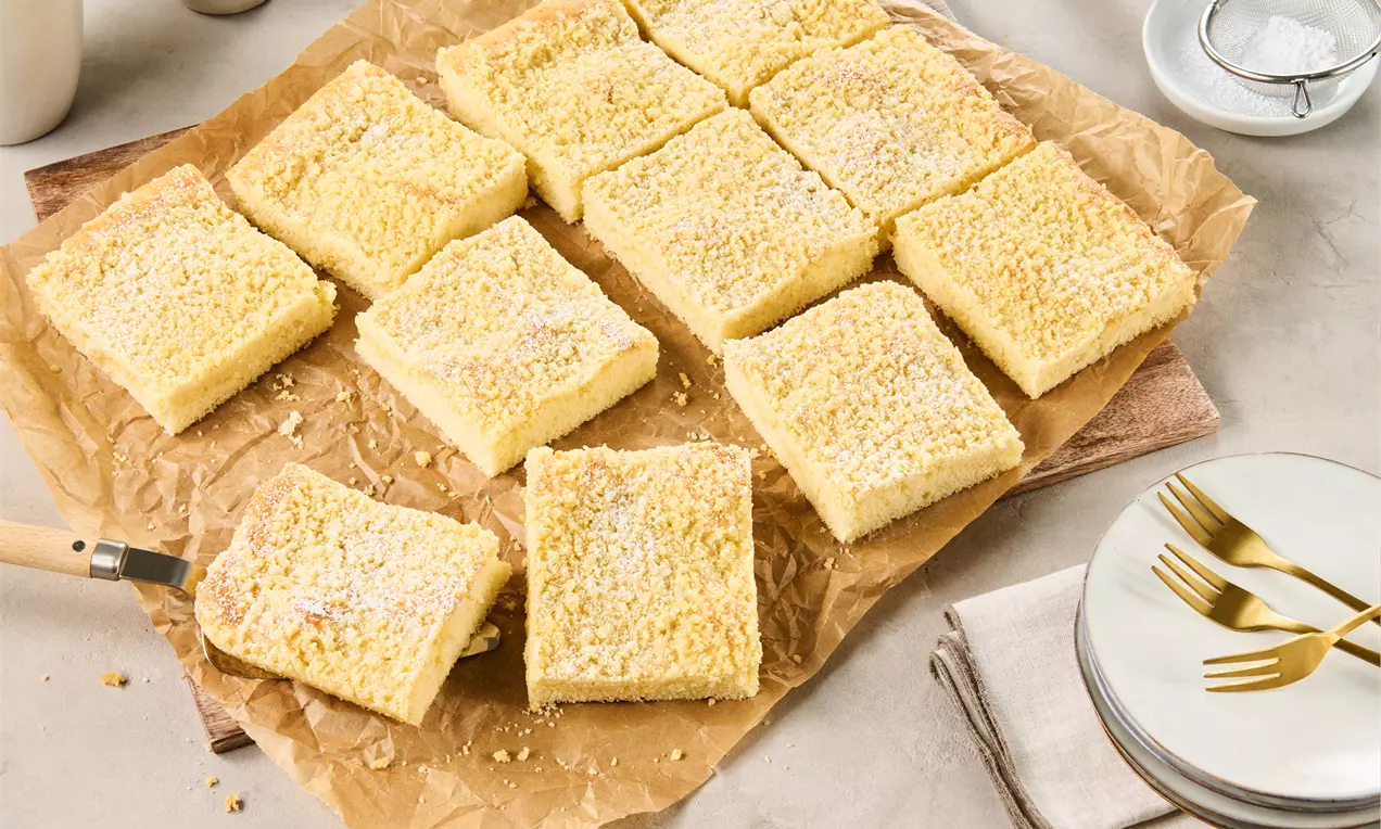 Quadratische Stücke Streuselkuchen mit goldgelber Kruste und feinen Streuseln auf braunem Backpapier, daneben Teller und Besteck