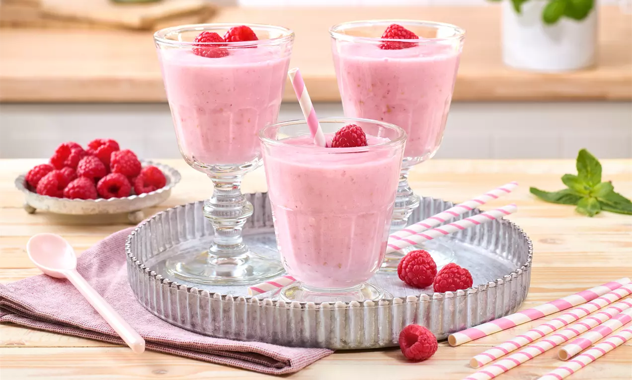 Cremiger, rosa Himbeer-Lassi in Gläsern mit Himbeerdekoration und gestreiften Trinkhalmen auf einem silbernen Tablett, umgeben von frischen Himbeeren