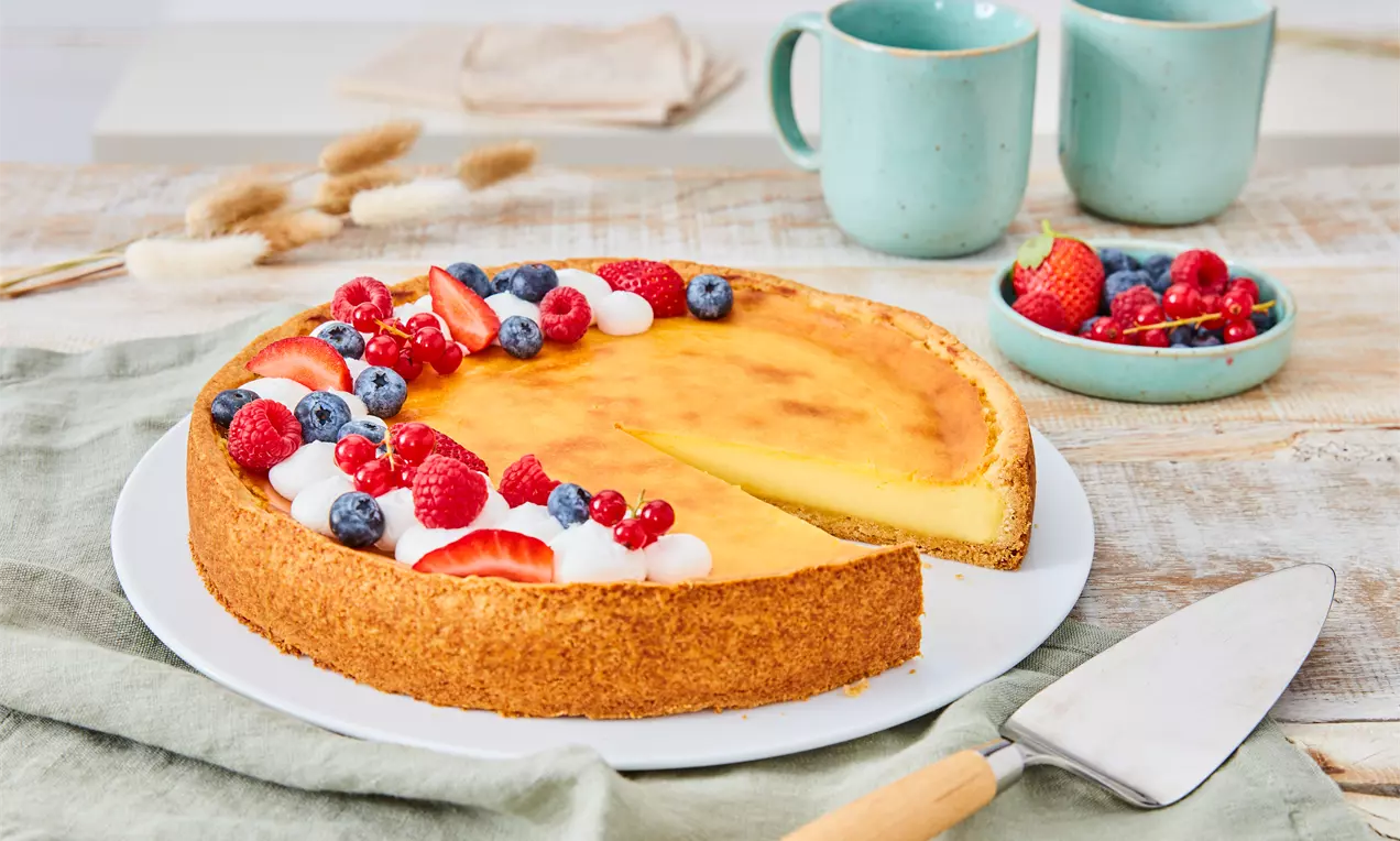 Veganer Käsekuchen mit goldgelber Oberfläche, auf weißem Teller präsentiert, garniert mit Sahnetupfen, roten und blauen Beeren