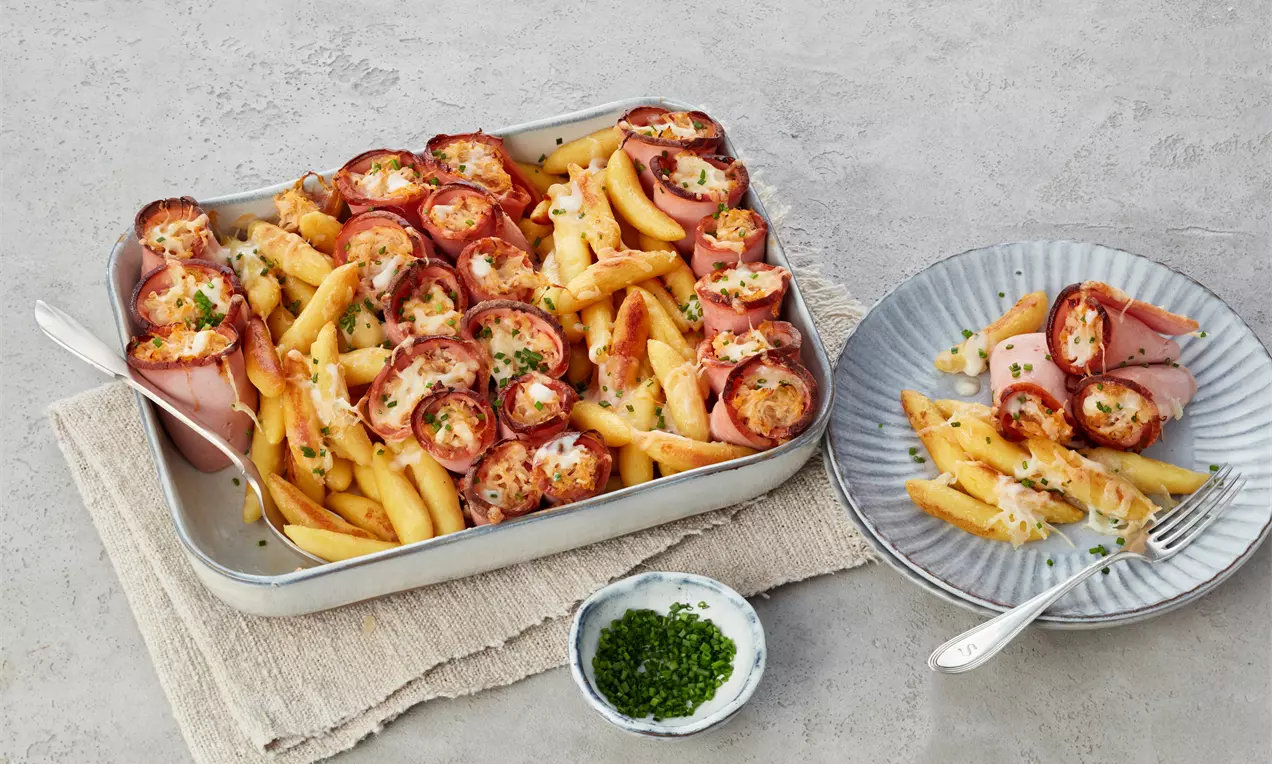Goldbraune Penne und mit Käse gefüllte Leberkäseröllchen, gebacken in einer silbernen Auflaufform, garniert mit frischen Schnittlauchringen