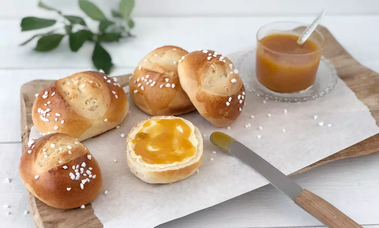 Goldene Camping-Brötchen mit Kristallzucker auf weißem Papier, ein halbiertes Brötchen bestrichen mit gelber Mirabellen-Konfitüre, Holzbrett und Glas