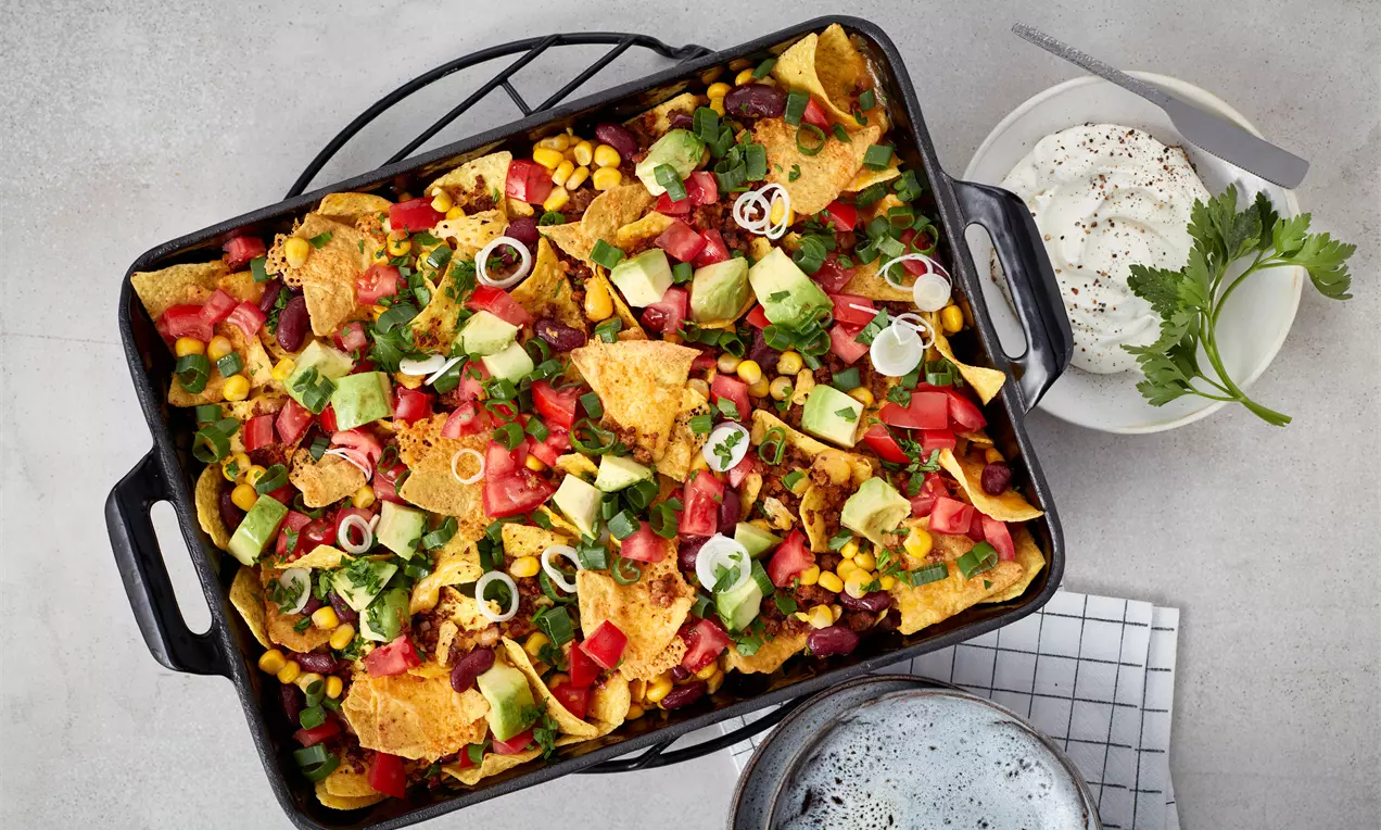 Bunte Mischung aus knusprigen Nachos, leuchtendem Gemüse, cremiger Avocado und frischen Kräutern in einer schwarzen Auflaufform