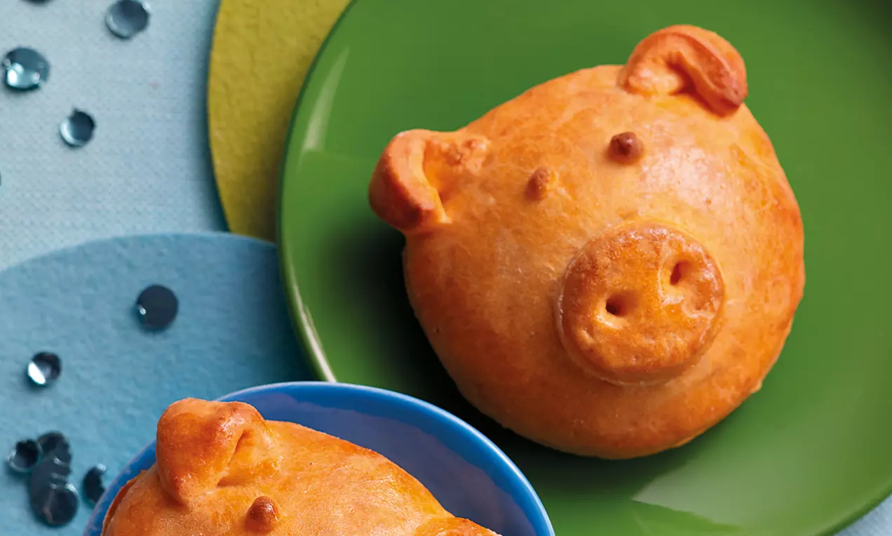 Goldbraune, rund geformte Hefeteig-Glücksschweinchen mit geprägtem Gesicht auf grünem Teller, daneben ein Schweinchen in blauer Schale