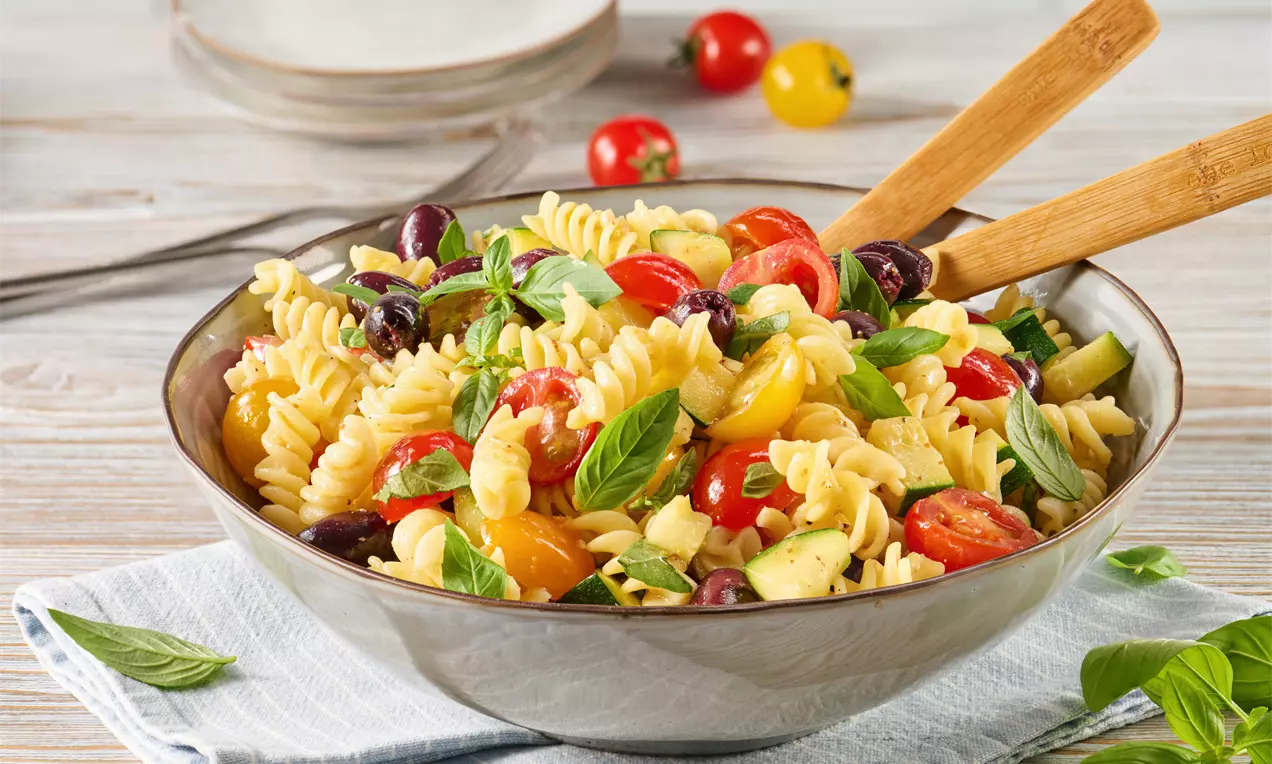 Bunter Nudelsalat aus goldenen Fusilli, roten und gelben Cherrytomaten, schwarzen Oliven und Basilikumblättern, angerichtet in einer Schüssel mit Holzsalatlöffeln