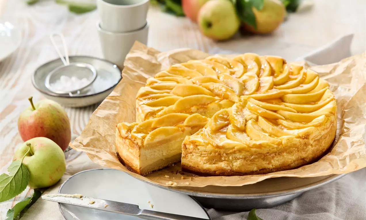 Goldbrauner Apfelkuchen mit glänzendem Belag aus dünnen Apfelscheiben, angerichtet auf Backpapier, umgeben von frischen Äpfeln und Geschirr