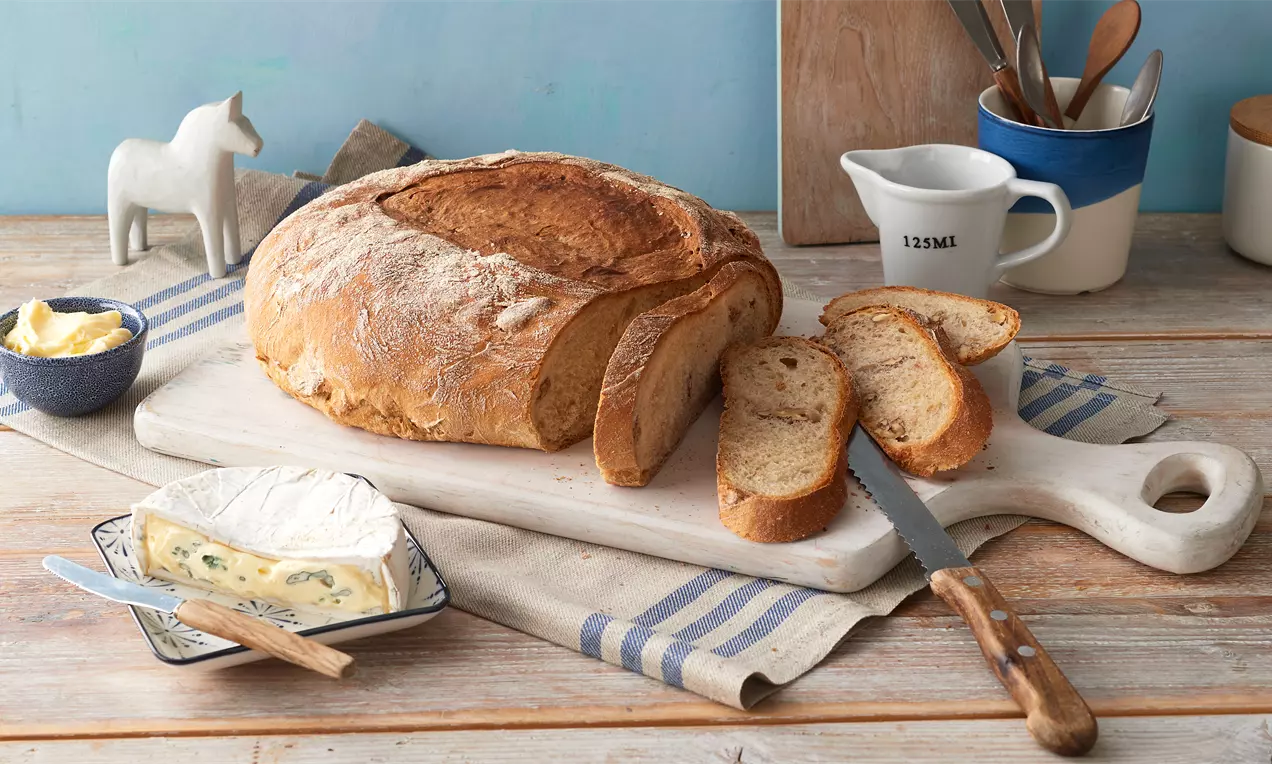 Rustikal gebackenes Walnussbrot mit goldbrauner Kruste, auf einem Holzbrett serviert, begleitet von Käse, Butter, Messer und Geschirr