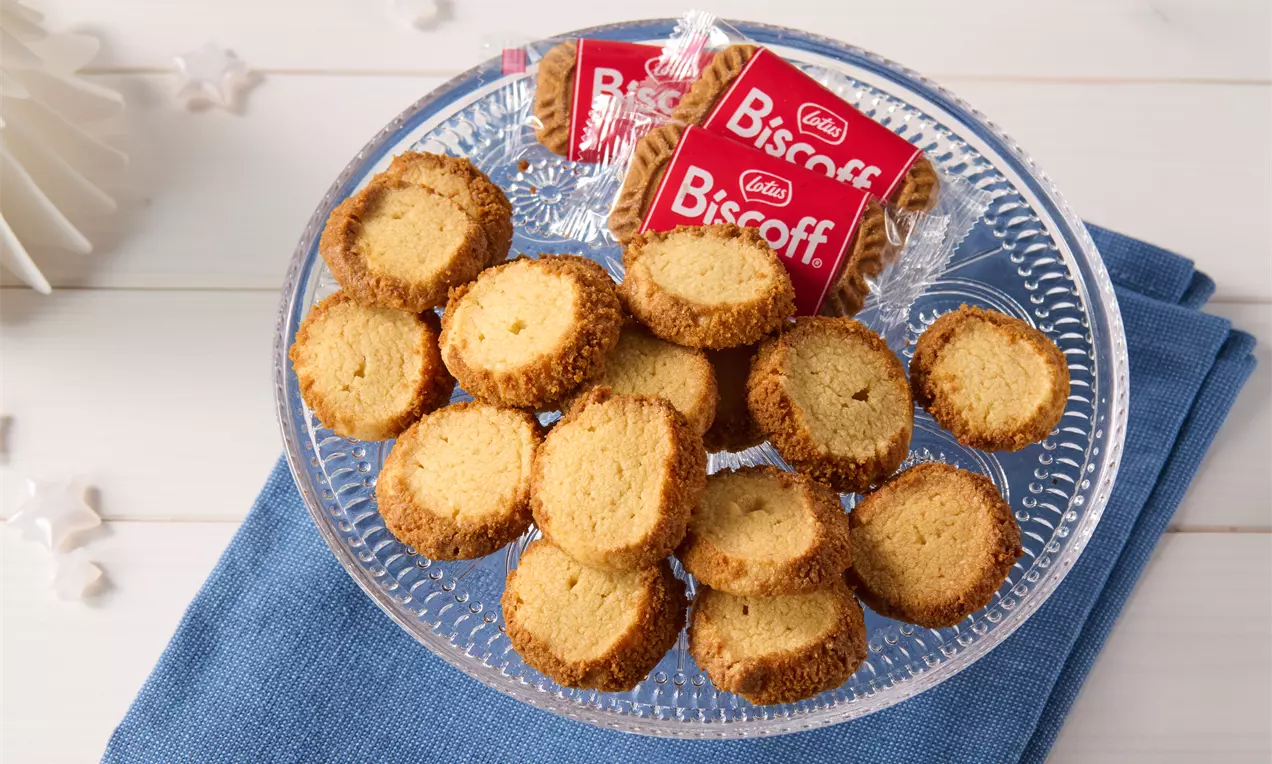 Knusprige, goldbraune Lotus-Kekstaler mit glatter Oberfläche und karamellfarbenem Rand auf einer gläsernen Platte, dekoriert mit verpackten Biscoff-Keksen