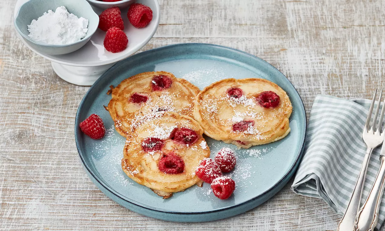 Goldene Pancakes mit eingebackenen Himbeeren, locker bestäubt mit Puderzucker, angerichtet auf einem blauen Teller neben einer Stoffserviette