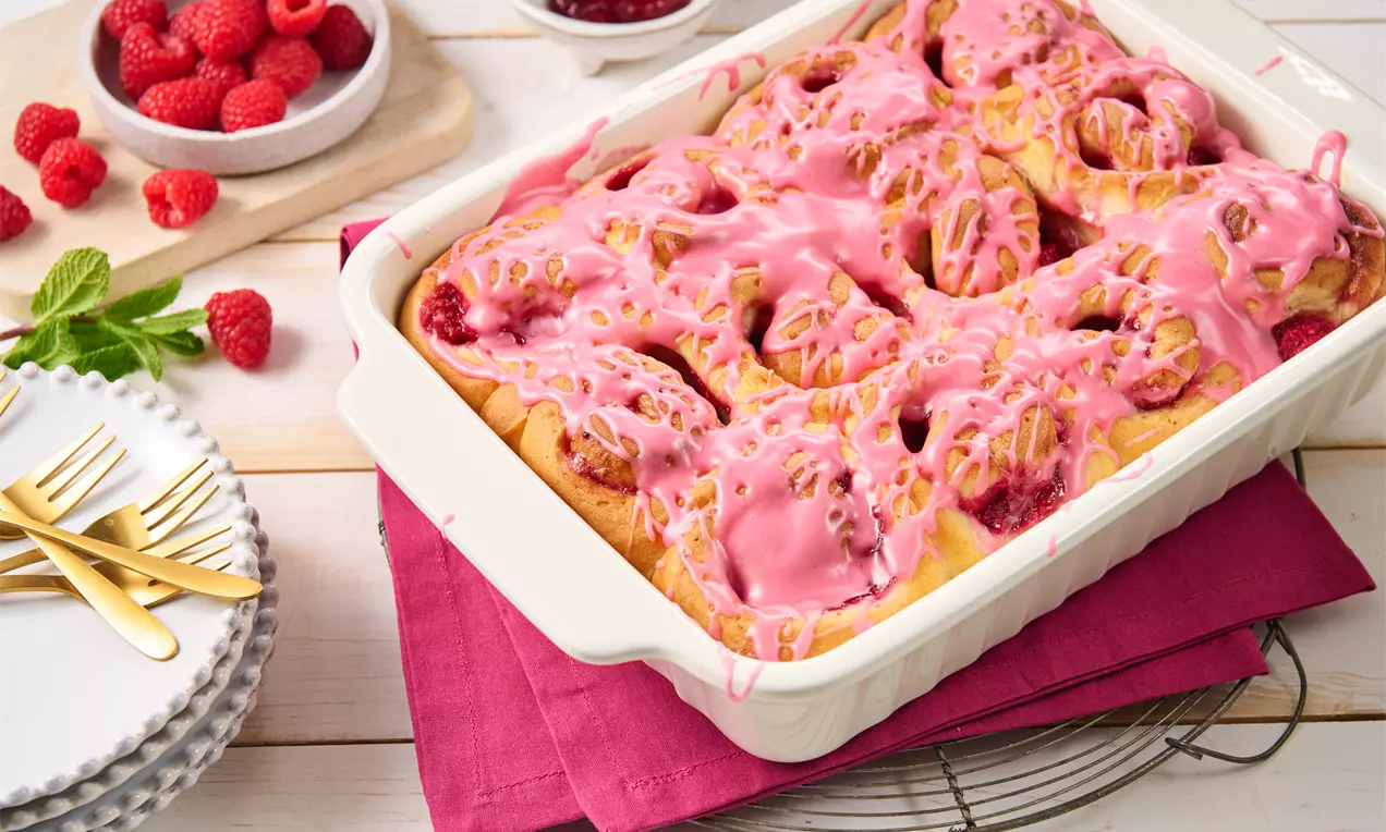 Frische Himbeer-Schnecken mit goldbraunem Teig und zartrosa Glasur, serviert in einer weißen Form auf pinkfarbenem Stoff