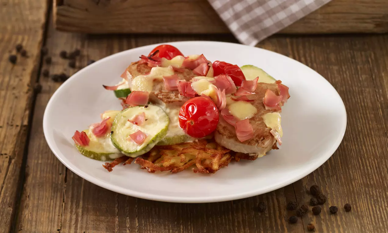 Überbackenes Schweinefilet mit heller Kruste auf Rösti, belegt mit geschmolzenem Käse, Schinkenwürfeln, Zucchinischeiben und roten Cherrytomaten