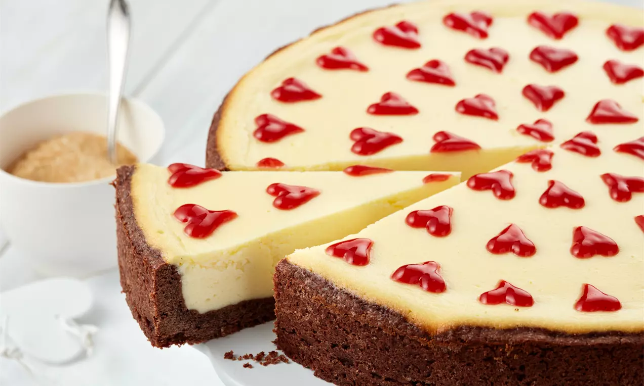Cremig-gelber Käsekuchen auf braunem Keksboden, verziert mit roten Herzchen aus Fruchtgelee, auf einer weißen Tortenplatte serviert