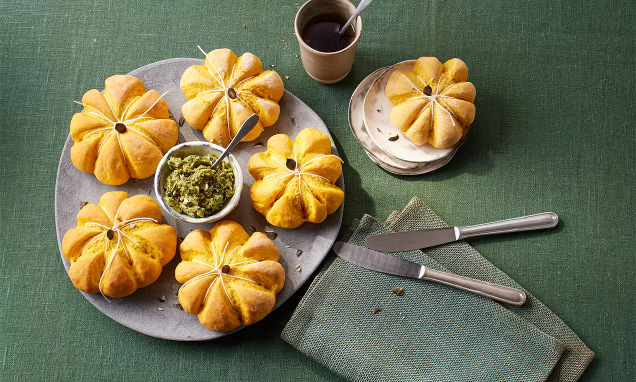 Goldorange Kürbisbrötchen mit bauchiger Form auf rundem Teller, dazu Schälchen mit grünem Aufstrich, Messer und Tasse auf grünem Tuch