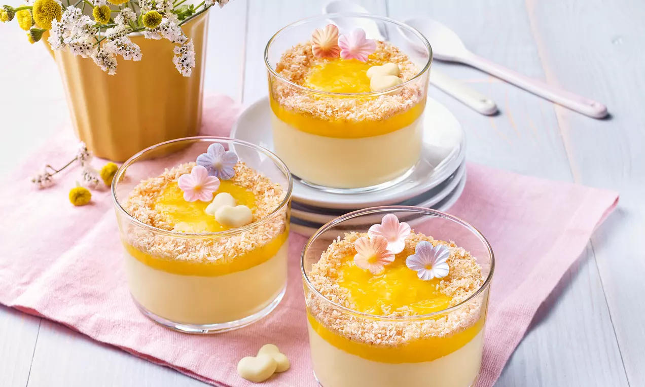Gläser mit cremigem Kokos-Pudding, leuchtend gelbem Mangopüree, gerösteten Kokosflocken und bunten Blüten auf rosa Stoff drapiert