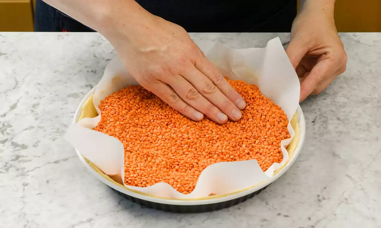 Mürbteigboden in runder Tarte-Form mit weißem Backpapier ausgelegt, rotem Blindbackgewicht aus Linsen bedeckt, auf heller Steinplatte