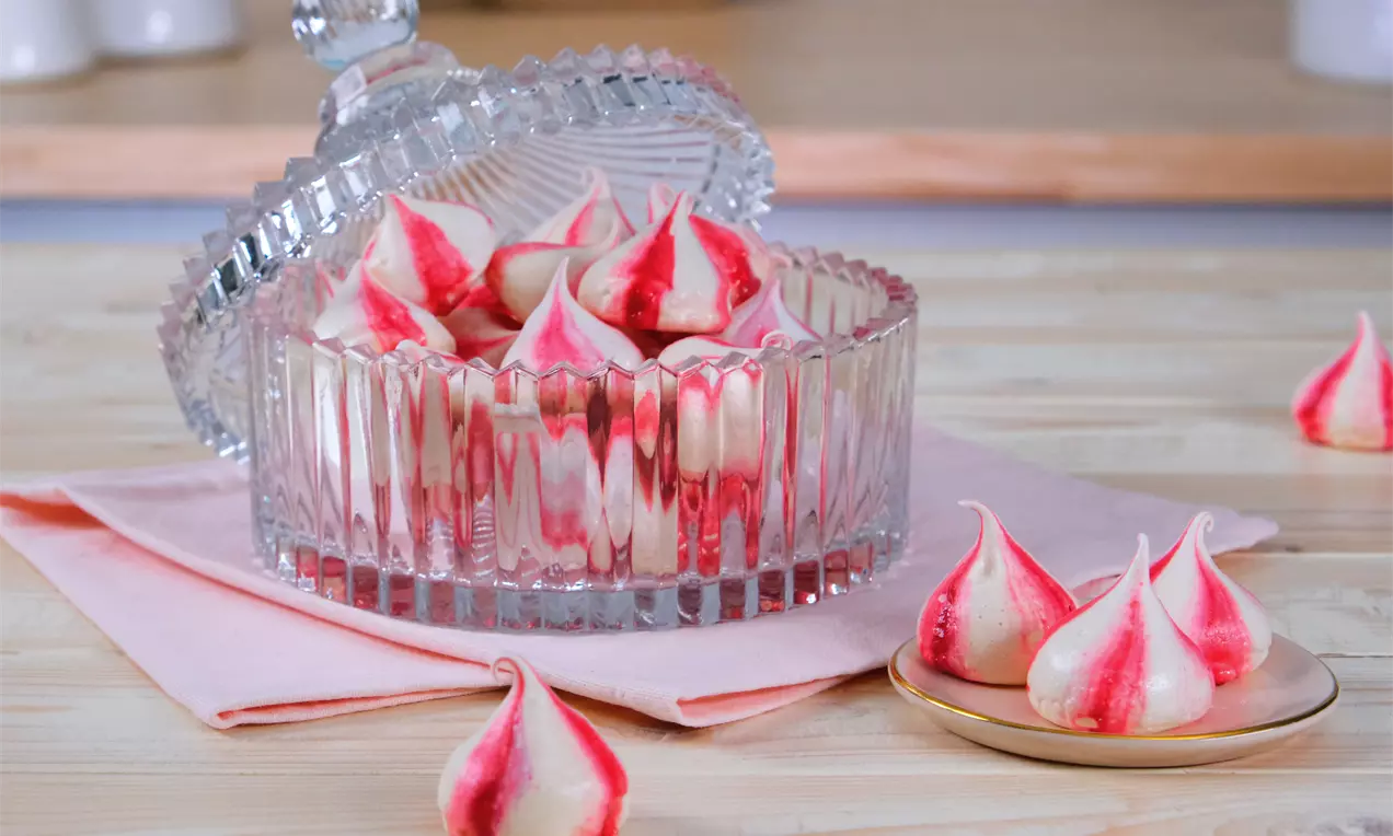 Zart rosa-weiß marmorierte Baiserküsschen in einer facettierten Kristallschale mit Glasdeckel, umgeben von einzelnen Küsschen auf Holz und Rosa