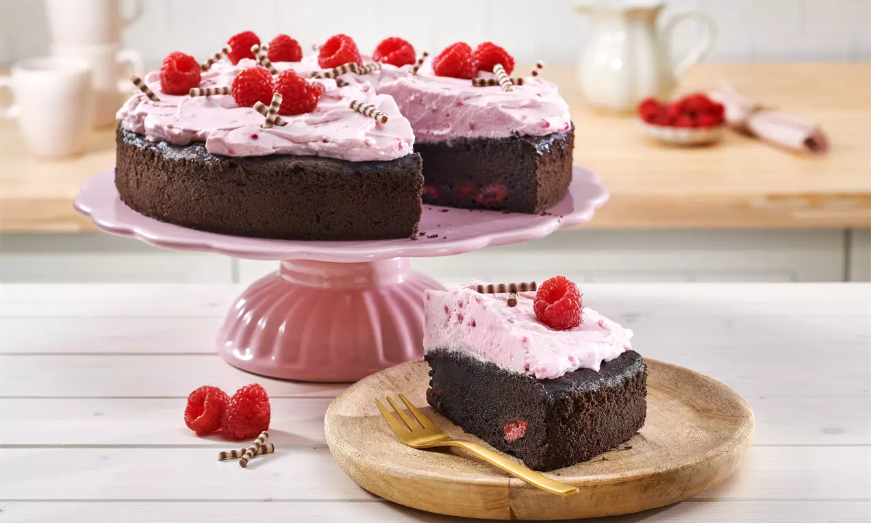 Schoko-Himbeer-Kuchen mit dunklem Boden, roséfarbener Creme und frischen Himbeeren auf rosa Platte und einem Stück auf Holzbrett mit Gabel