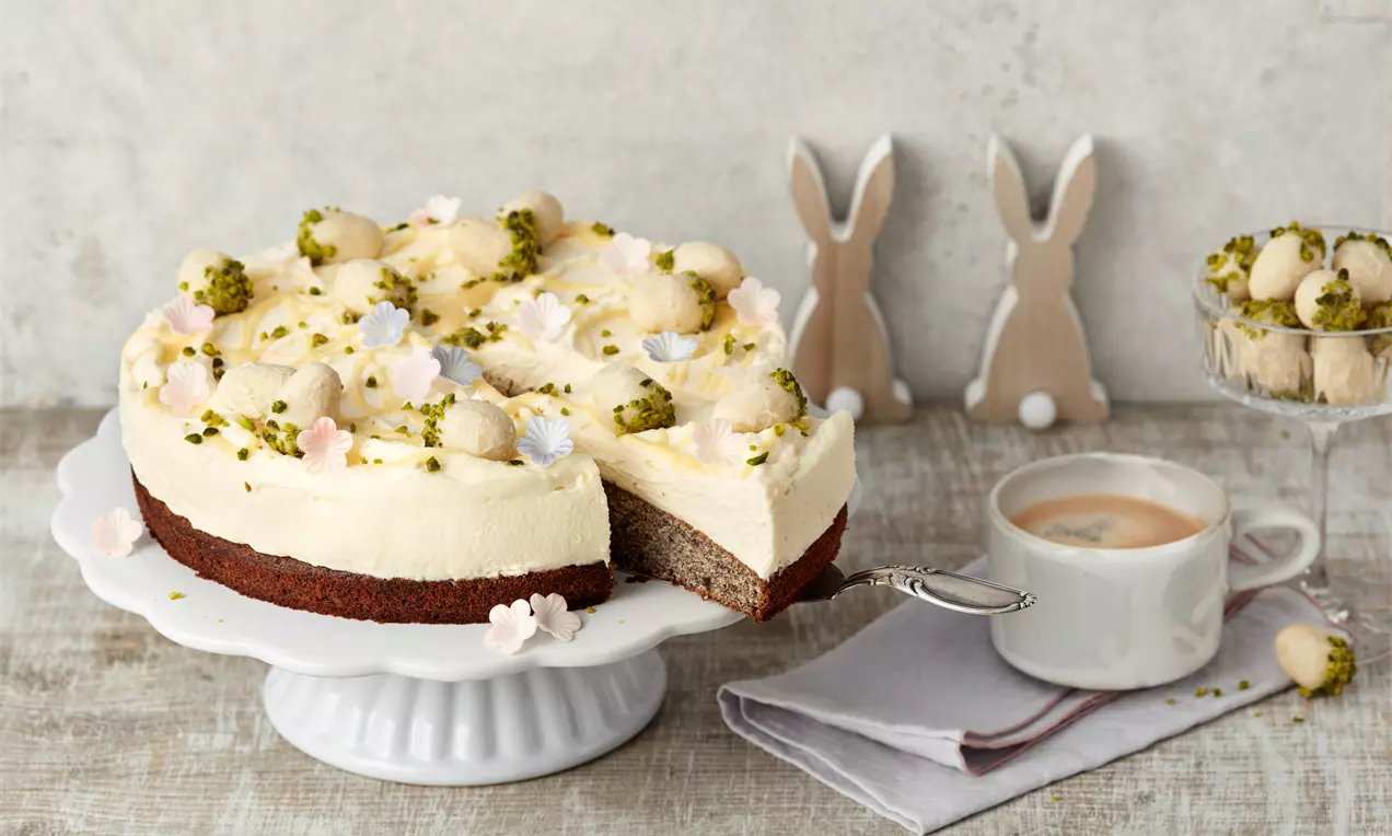 Eierlikör-Mohntorte mit heller Creme, gelben Tupfen, Moosgrün-Pistazien und Blüten auf dunklem Boden auf weißer Tortenplatte