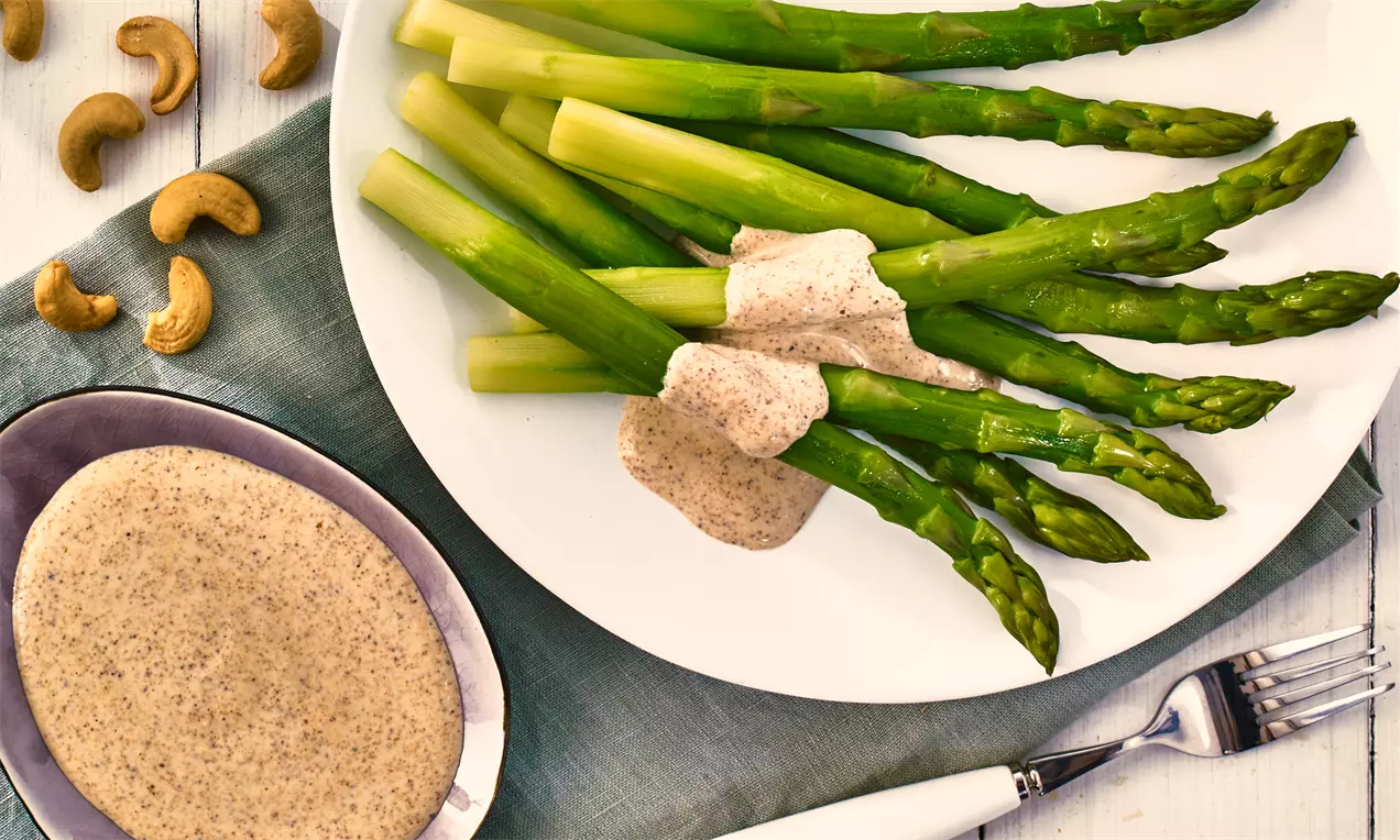Grüne Spargelstangen mit cremiger Cashew-Soße auf weißem Teller, daneben Schüssel mit Soße, Cashewkerne und graues Tuch mit Besteck