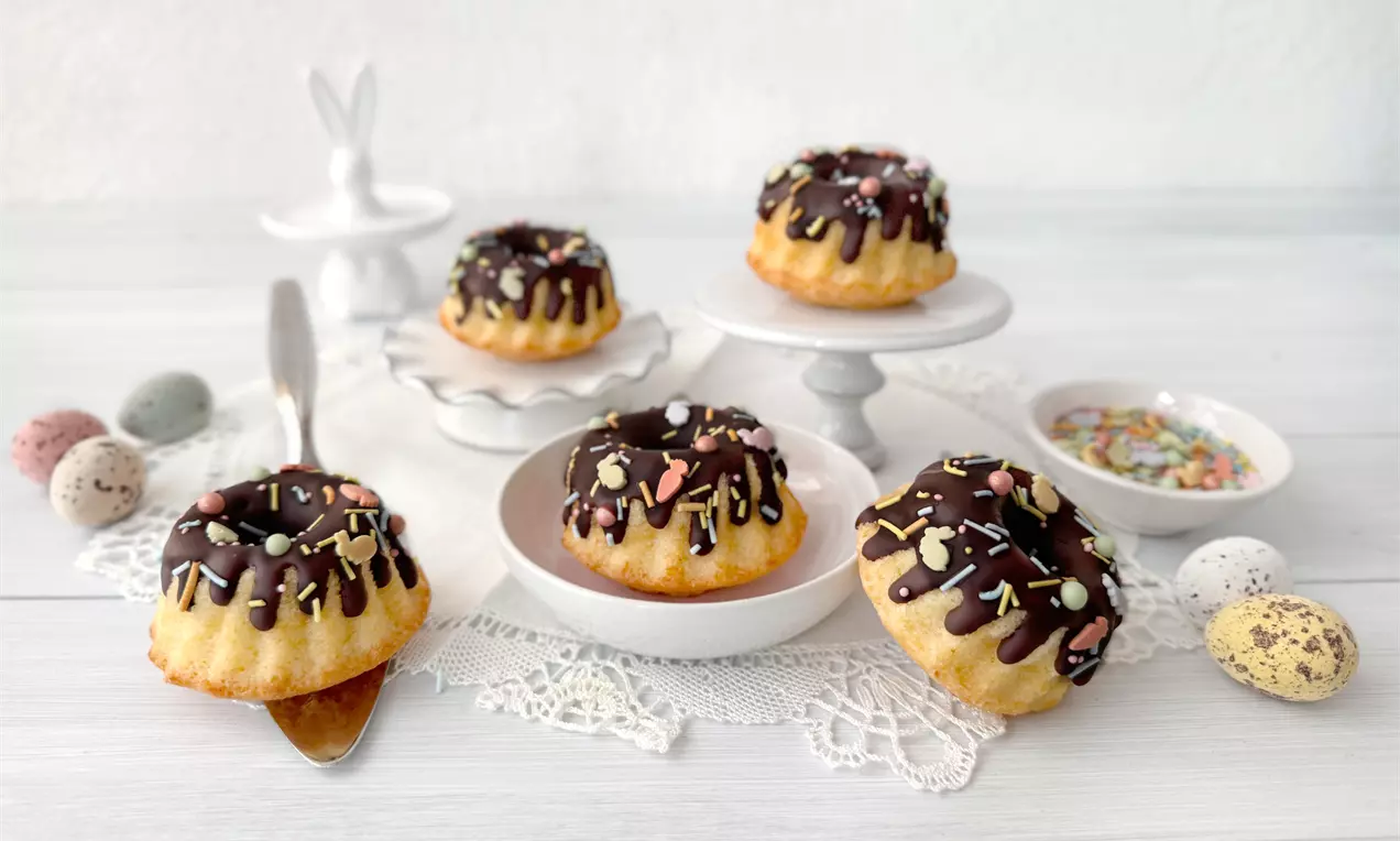 Mini-Gugelhupfe mit Eierlikör, goldgelb, mit glänzendem Schokoladenguss und pastellfarbener Osterdekoration auf weißem Teller