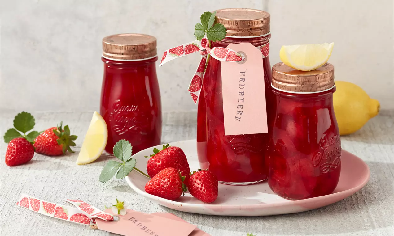 Gläser mit glänzend roter Erdbeer-Konfitüre, verziert mit Schleifen und Etiketten, arrangiert mit frischen Erdbeeren und Zitronenscheibe auf rosa Teller
