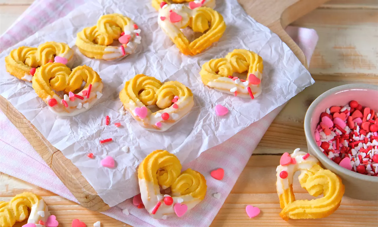 Spritzgebäck in Herzform, goldgelb mit weißer Glasur und rosa Zuckerstreuseln auf Pergamentpapier, daneben eine Schale mit Streudekor
