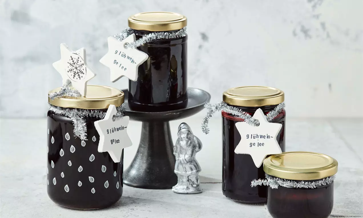 Vier Gläser mit tiefrotem Glühwein-Gelee, teils auf dunkler Etagere arrangiert, mit goldenen Deckeln und weißen Sternanhängern verziert